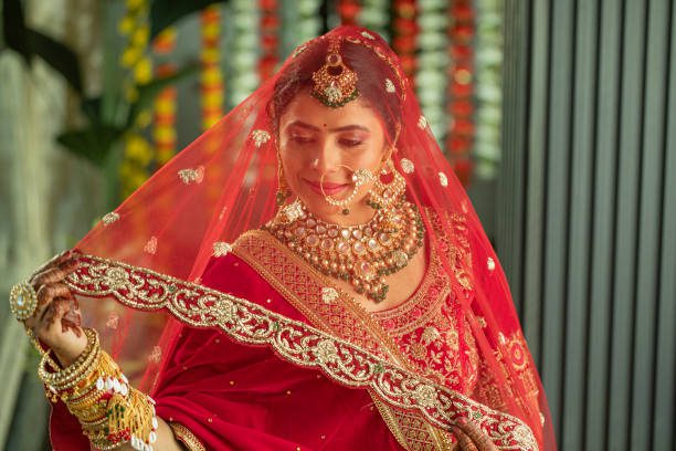 Portrait of beautiful bride dressed in traditional clothing and jewelries smiling and looking through red dupatta during wedding ceremony